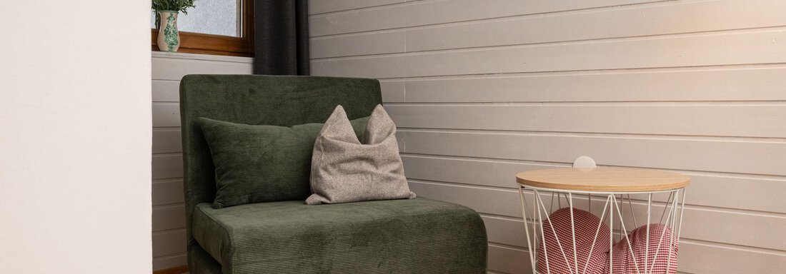A cozy corner with a green armchair and a side table. The room has bright walls and a window with curtains. - Ferienhaus Valsora - Sölden