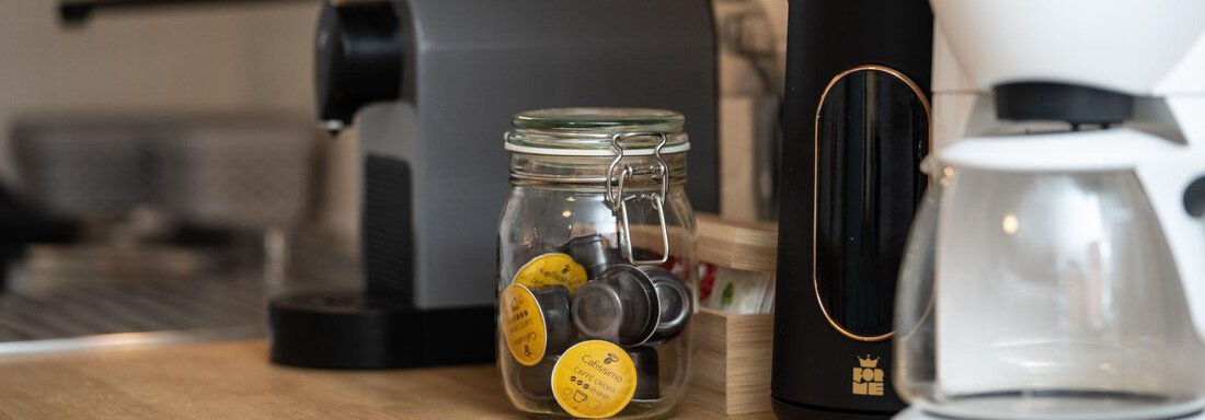 A modern kitchen unit with various appliances. A glass with coffee capsules is placed next to a coffee machine and a kettle on a wooden table. - Ferienhaus Valsora - Sölden