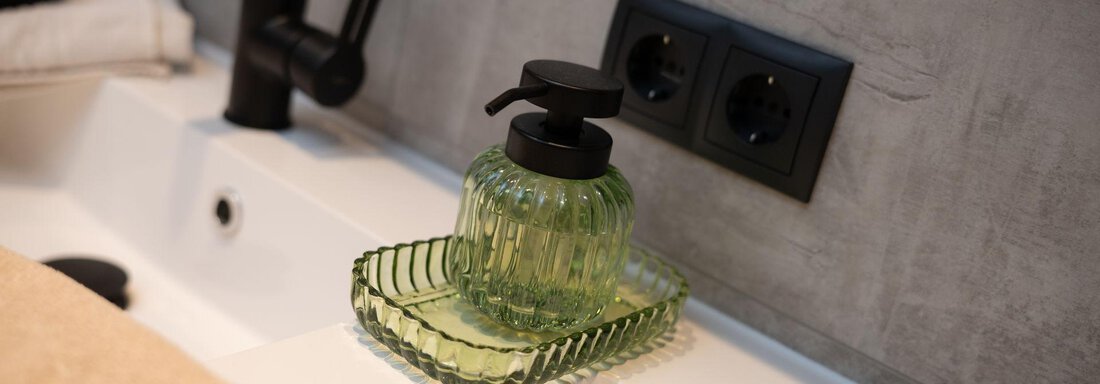 An elegant soap dispenser made of green glass is on a sink. Next to it lies a towel in a neutral color. - Ferienhaus Valsora - Sölden