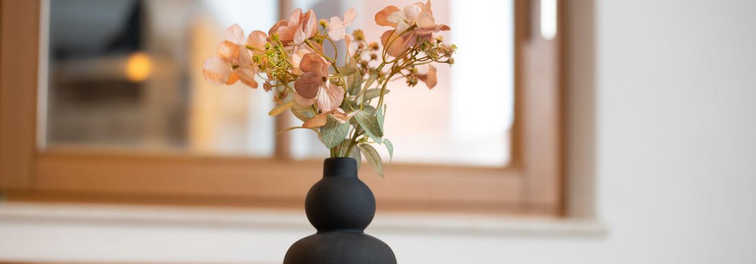 A stylish black vase with delicate pink flowers stands on a wooden table. In the background, a window with natural light can be seen. - Ferienhaus Valsora - Sölden