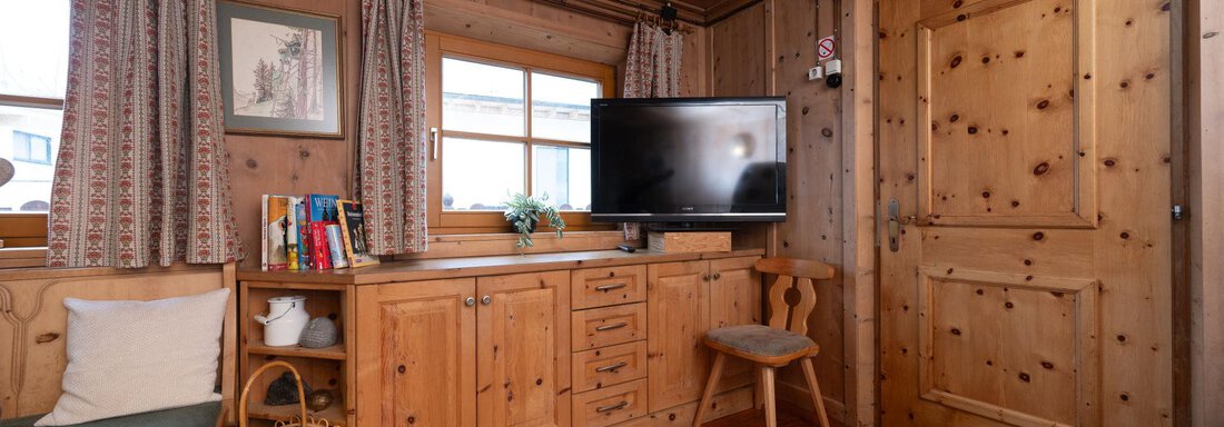 A cozy room with wood paneling and large windows. There is a TV, a desk, and a seating area. - Ferienhaus Valsora - Sölden