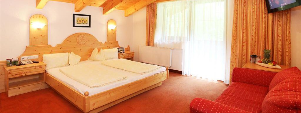 A cozy bedroom with wooden decor, a large bed, and a red sofa. The room has large windows that let in plenty of light. - Ferienhaus Waldele - Sölden