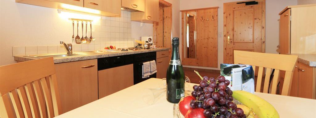 A modern kitchen with wooden shelves and a dining table. On the table sits a fruit bowl with grapes, apples, and bananas, as well as a bottle of champagne. - Ferienhaus Waldele - Sölden