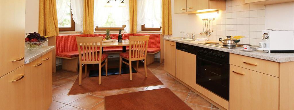 A modern kitchen with wooden cabinets and a dining area. The room has large windows and a cozy atmosphere. - Ferienhaus Waldele - Sölden