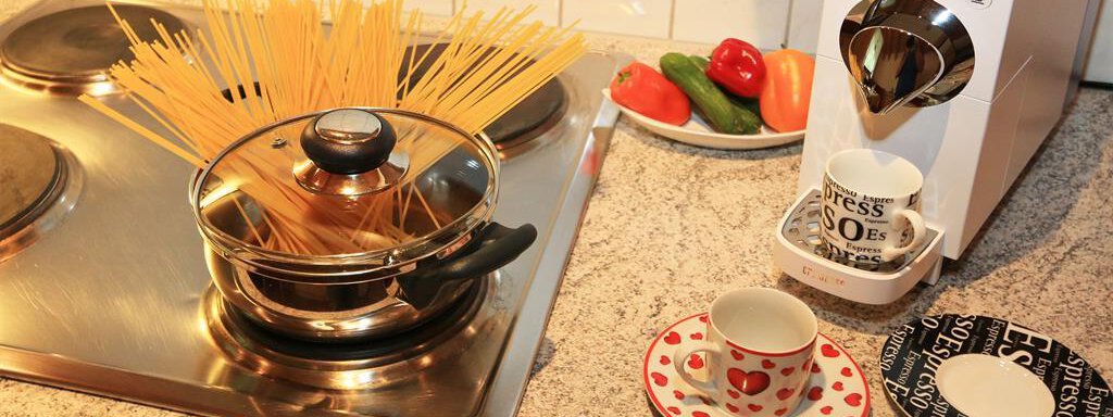 A pot with spaghetti on a stove is next to a coffee machine. On the table, there are two cups, a bowl with vegetables, and a decorated saucer. - Ferienhaus Waldele - Sölden
