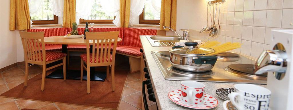 A cozy kitchen with a dining table and a bench. There are bright curtains and modern kitchen equipment. - Ferienhaus Waldele - Sölden