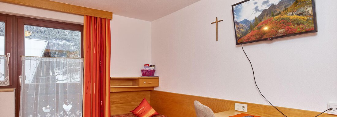 A simple room with a single bed, a desk, and a TV on the wall.  
Large windows provide access to a balcony with a view. - Ferienhof Paulelas - Sölden