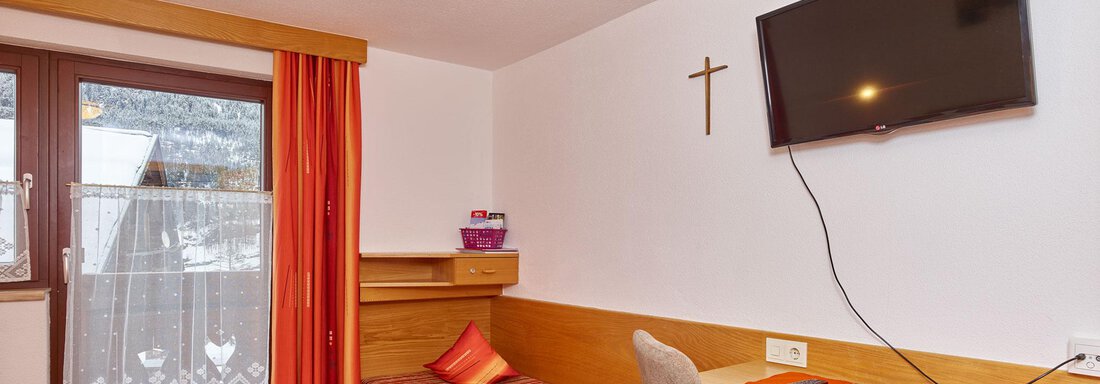A simple single room with a bed, a desk, and a television. The windows have red curtains and offer a view outside. - Ferienhof Paulelas - Sölden