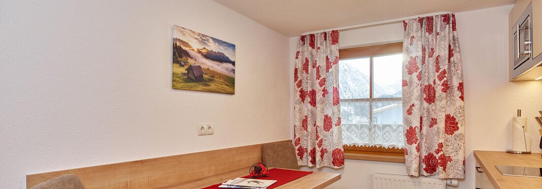A cozy dining area with a table and two chairs. The walls are painted in light colors and adorned with a landscape photograph and floral curtains. - Ferienhof Paulelas - Sölden