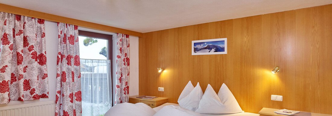 A cozy bedroom with a large bed and wood paneling. The windows are decorated with colorful curtains and there is a beautiful view. - Ferienhof Paulelas - Sölden