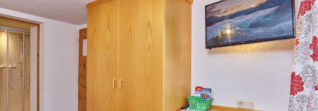 A simple room with a wooden wardrobe, a desk, and a large window with curtains. On the wall hangs a picture of a landscape. - Ferienhof Paulelas - Sölden