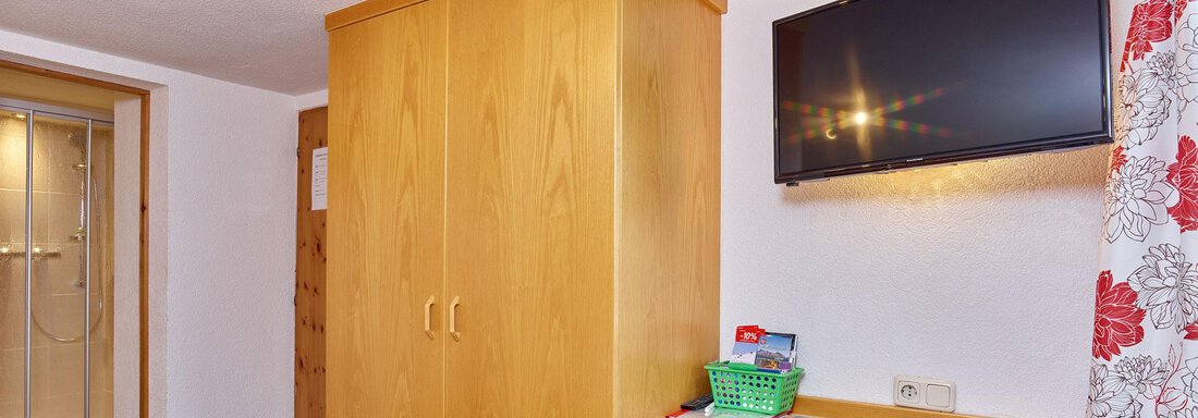 A simple room with a wooden wardrobe, a desk, and a TV on the wall. The room has a pleasant workspace and a bright atmosphere. - Ferienhof Paulelas - Sölden