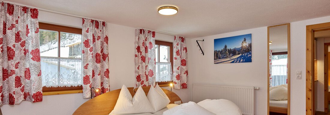 A cozy bedroom with a large double bed and red carpets. Flower-patterned curtains and a beautiful view of the mountains create an inviting atmosphere. - Ferienhof Paulelas - Sölden