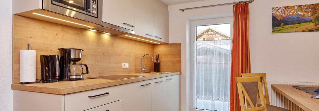 A modern kitchen with wooden surfaces and a microwave. A dining table with chairs and a window with curtains are also visible. - Ferienhof Paulelas - Sölden