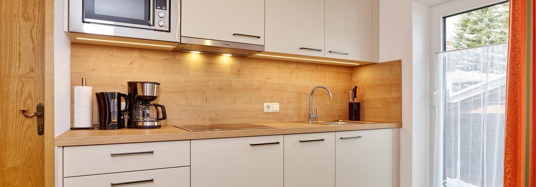 A modern kitchen with light cabinets and a wooden countertop. There is a kettle, a coffee maker, and a window with an orange curtain. - Ferienhof Paulelas - Sölden
