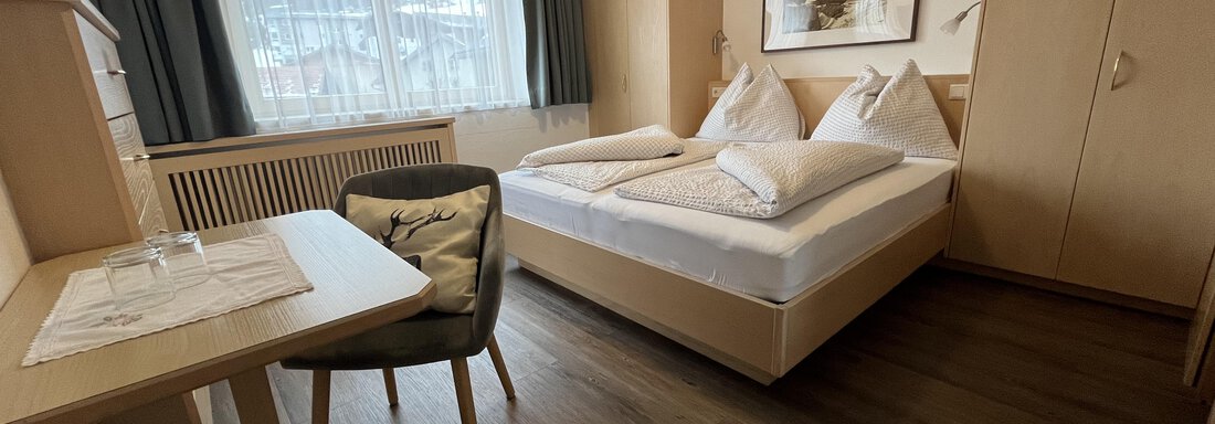 A cozy room with a double bed, desk, and chair. Large windows provide plenty of daylight. - Franzele Appartements & Zimmer - Sölden