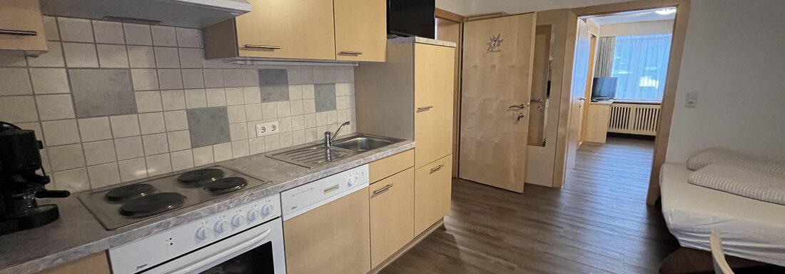 A modern kitchen with wooden cabinets and white appliances. In the background, a bright living area is visible. - Franzele Appartements & Zimmer - Sölden