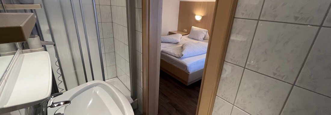 A modern bathroom with a shower and a sink. In the background, a bedroom with a double bed can be seen. - Franzele Appartements & Zimmer - Sölden
