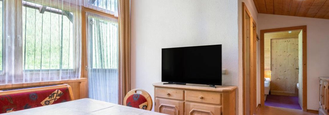 A cozy dining area with a table and chairs. In the corner stands a television, and large windows provide plenty of daylight. - Garni Nadine - Sölden