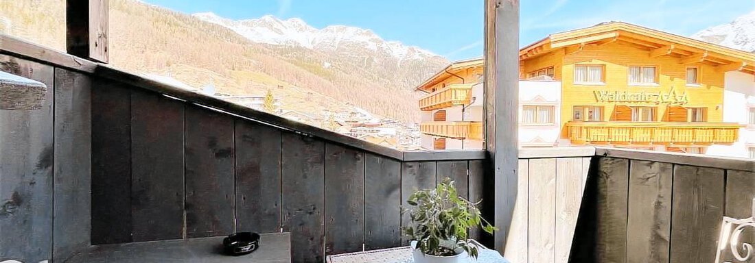 A cozy balcony with a table and chairs, surrounded by wood paneling. In the background, mountains and a bright blue sky can be seen. - Garni Nadine - Sölden
