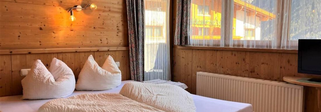 A cozy room with wooden walls and a large bed. There are soft pillows, a big window, and a television. - Garni Nadine - Sölden