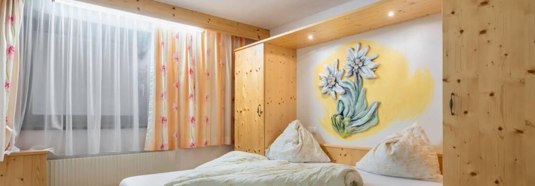 A cozy bedroom with wooden furniture and a large window. On the wall, there is a beautiful flower painting. - Garni Nadine - Sölden
