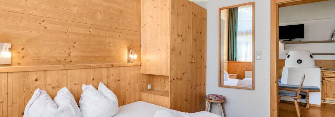 A cozy bedroom with wooden paneling and a large bed. The room is bright and inviting, with a small table and a mirror. - Garni Nadine - Sölden