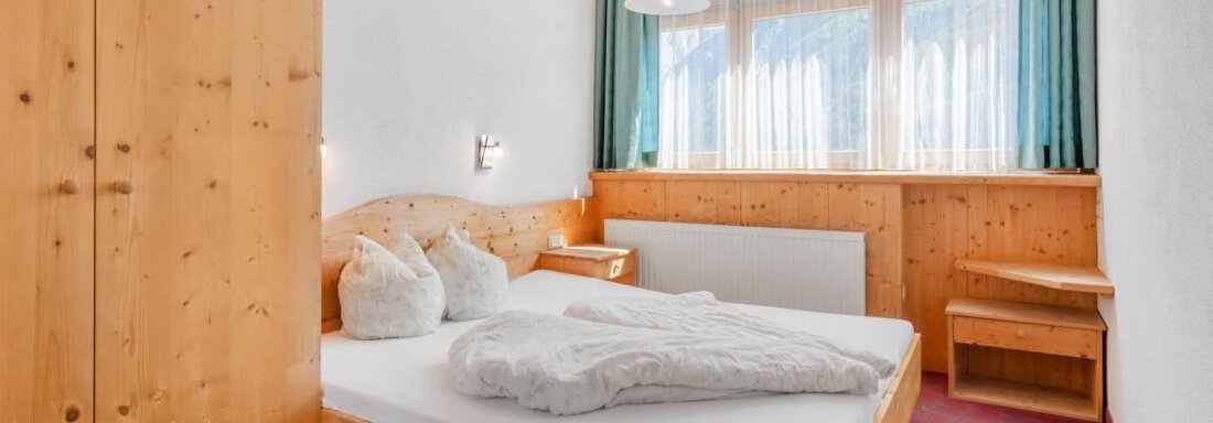 A cozy bedroom with wooden furniture and a large window. The room is bright and cheerful with a double bed and a small table. - Garni Nadine - Sölden