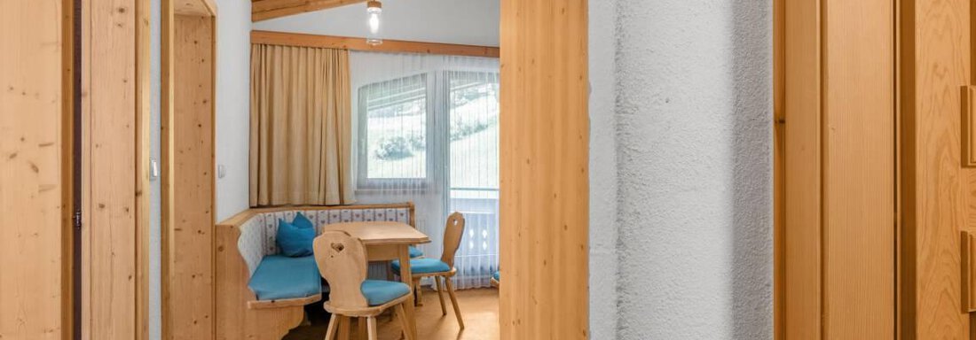 A bright hallway with wooden paneling and a corner with a table and chairs. Behind a window, there is a view of nature. - Garni Nadine - Sölden