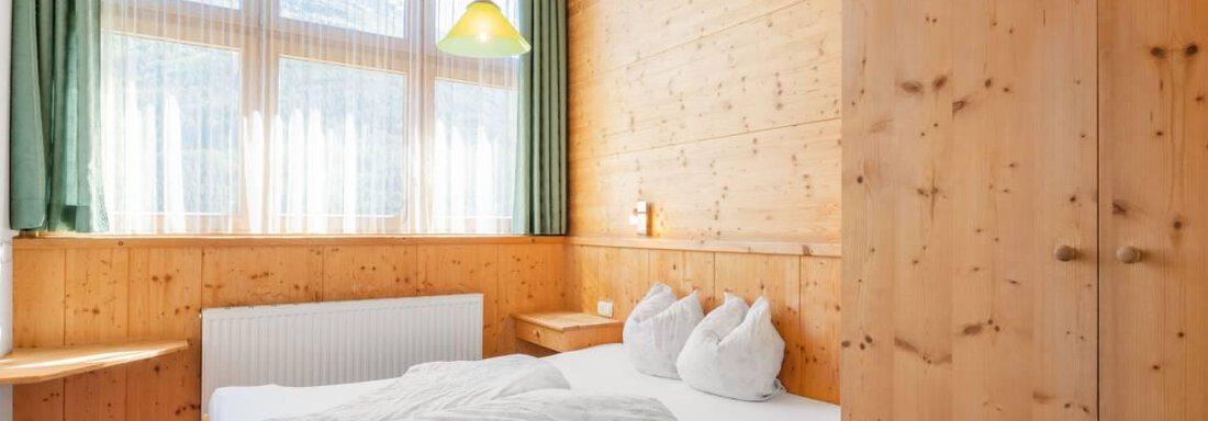 A cozy bedroom made of wood with a large window and plenty of daylight. The bed is outfitted with a white blanket and pillows. - Garni Nadine - Sölden