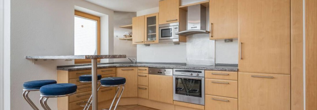 A modern kitchen with wooden cabinets and an elegant bar with blue stools. The simple furnishings create an open and friendly atmosphere. - Garni Nadine - Sölden