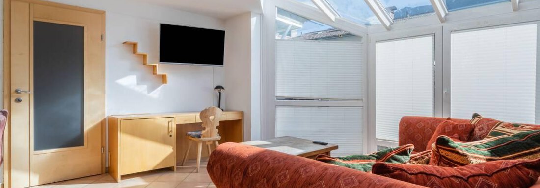 A bright living space with a large window and a red sofa. Also visible are a wooden shelf and a television on the wall. - Garni Nadine - Sölden