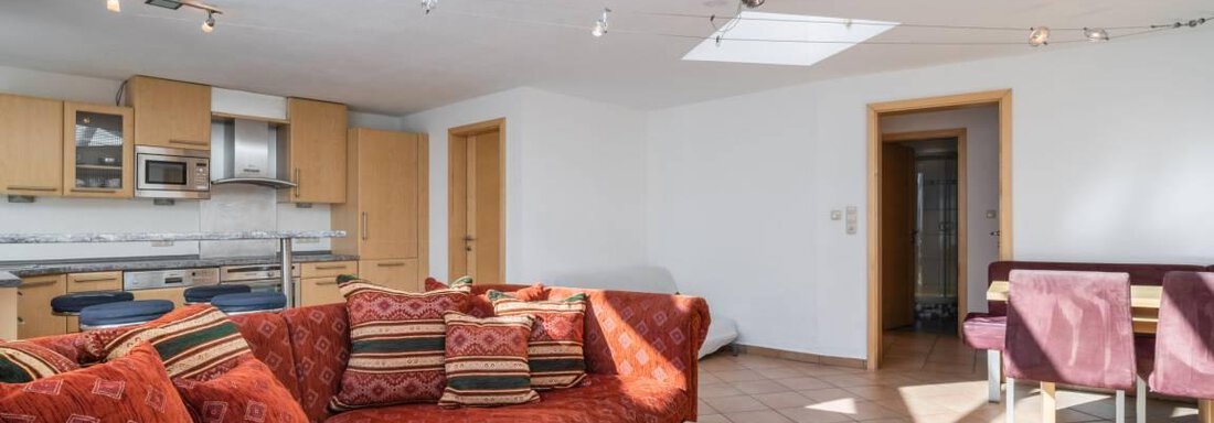 A bright living room with a red sofa and a wooden table. The open kitchen is well-equipped and there are large windows that let in a lot of light. - Garni Nadine - Sölden