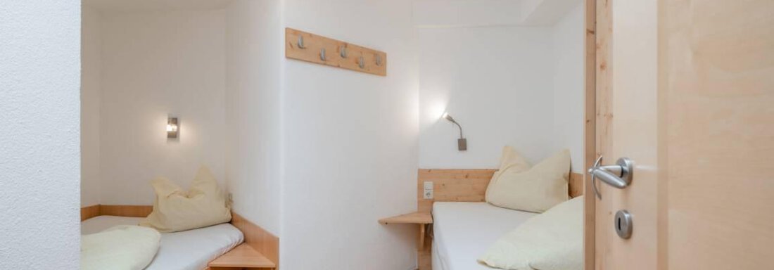 A simple room with two single beds and wooden details. The walls are light, and there is a modern ceiling light. - Garni Nadine - Sölden