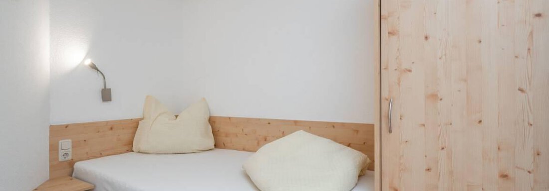 A simple room with bright walls and a single bed. There is a wooden shelf and a wardrobe, as well as cozy cushions. - Garni Nadine - Sölden