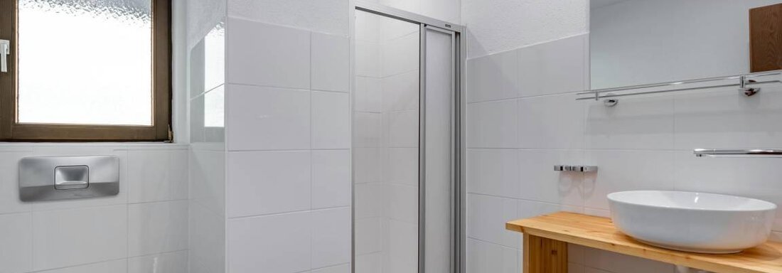 A modern bathroom with white tiles and a shower. A sink on a wooden shelf and a window provide bright lighting conditions. - Garni Nadine - Sölden