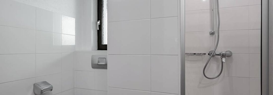 A modern bathroom with a shower and a toilet. The walls are designed with white tiles and there is a window for daylight. - Garni Nadine - Sölden