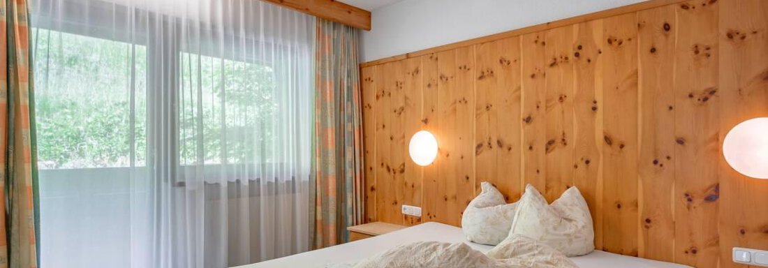 A cozy bedroom with a large bed and warm wooden walls. Large windows provide plenty of daylight and a view outside. - Garni Nadine - Sölden