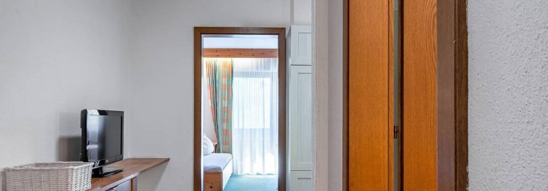 A cozy hallway with a wooden ceiling and a television. In the background, a bright room with a bed and a balcony is visible. - Garni Nadine - Sölden