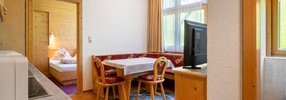 A cozy apartment with wood paneling and an open living area. The space features a dining table, a television, and a connection to a sleeping area. - Garni Nadine - Sölden