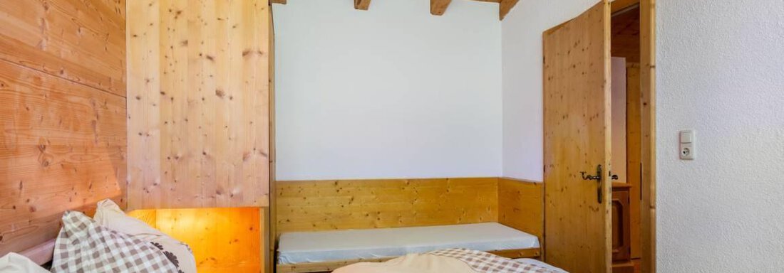 A cozy bedroom with wooden walls and a warm atmosphere. The room features a bed with checkered pillows and an additional sleeping option. - Garni Nadine - Sölden