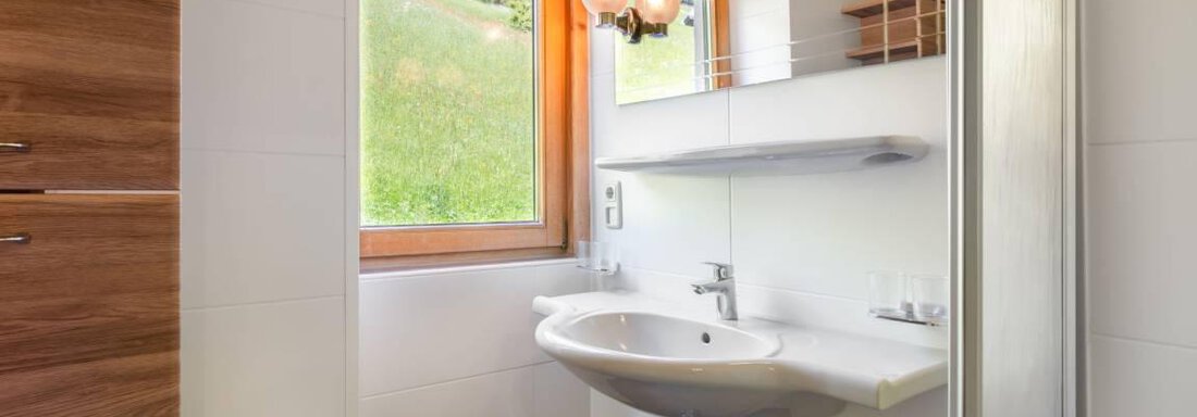 A modern bathroom with a sink and a mirror.  
Large windows provide plenty of natural light and a view of the greenery. - Garni Nadine - Sölden