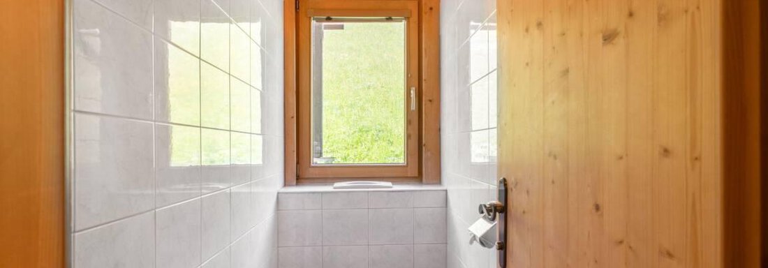 A modern bathroom with white tiles and a window. The wooden door and frame give the room a warm atmosphere. - Garni Nadine - Sölden