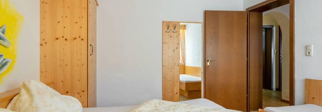 A cozy bedroom with wooden paneling and warm lighting. There are two beds and an open door leading to another room. - Garni Nadine - Sölden