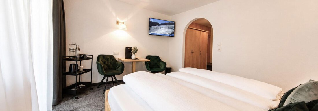 A modern hotel room with two beds and a cozy seating area. There is a table, a television, and large windows that let in plenty of light. - Garni Rustica - Sölden