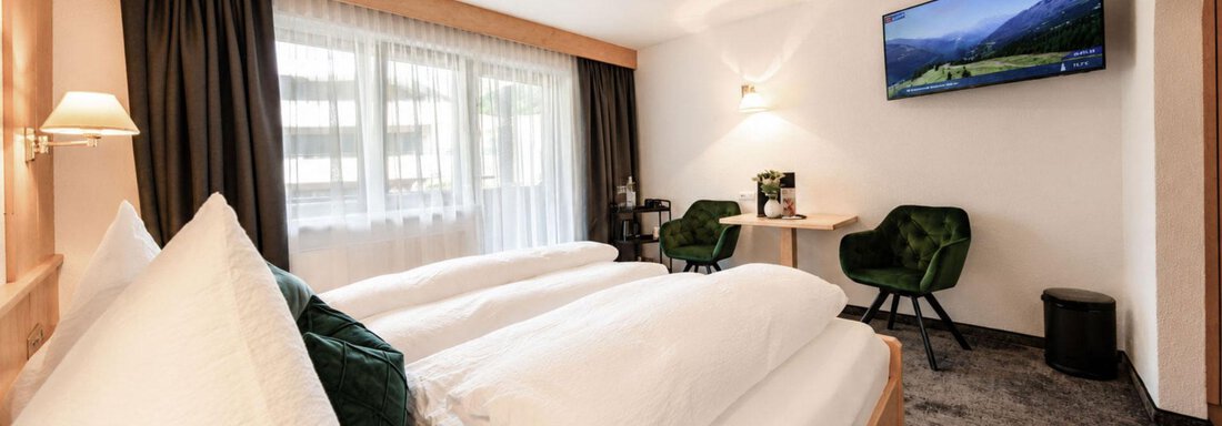 A modern hotel room with two cozy beds and a television. There is a small seating area with two green chairs and plenty of daylight. - Garni Rustica - Sölden