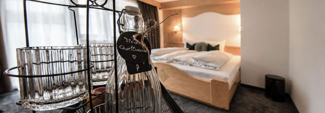 A modern room with a cozy bed and bright windows. In the foreground, there is a glass carafe and glasses on a tray. - Garni Rustica - Sölden
