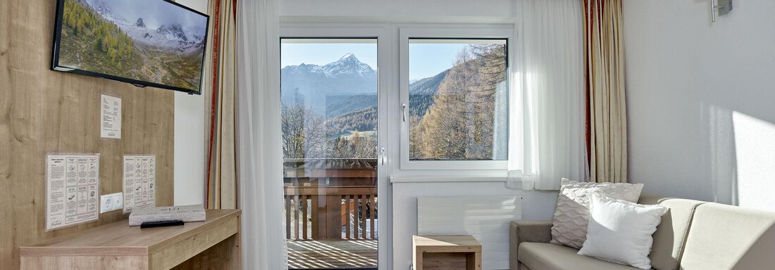 A bright living room with a sofa and a television. The windows offer a view of the mountains. - Gästehaus Bergkristall - Sölden