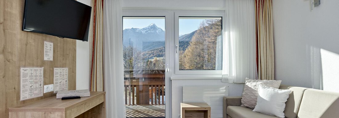 A cozy living room with a modern sofa and a television. Large windows offer a view of the mountains and plenty of natural light. - Gästehaus Bergkristall - Sölden