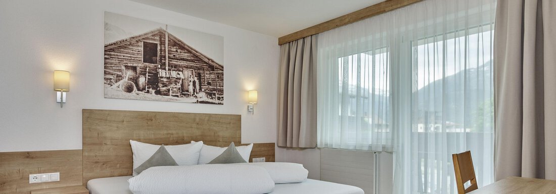 A modern bedroom with a wooden frame bed and soft colors. Large windows let in plenty of light and offer a view outside. - Gästehaus Bergkristall - Sölden